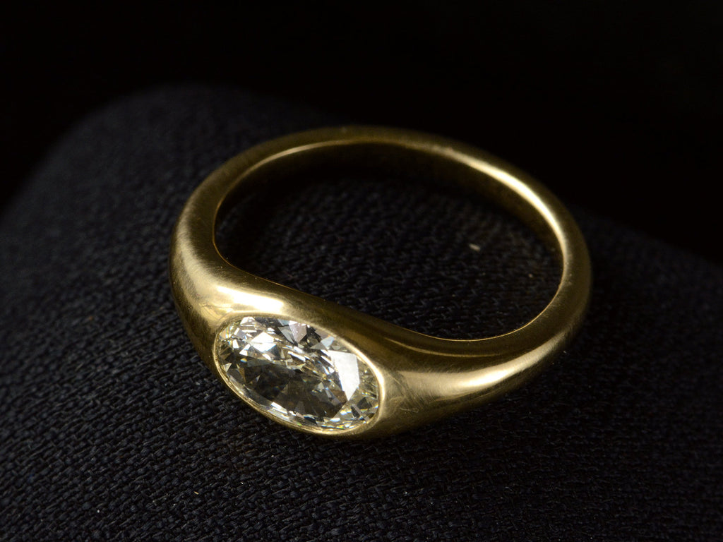 Elevated angled view of flush set oval diamond ring in yellow gold, shown on dark background.