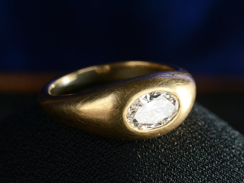 Elevated angled view of EB Modern flush set oval white diamond in rounded 22K yellow gold setting, shown on dark background.