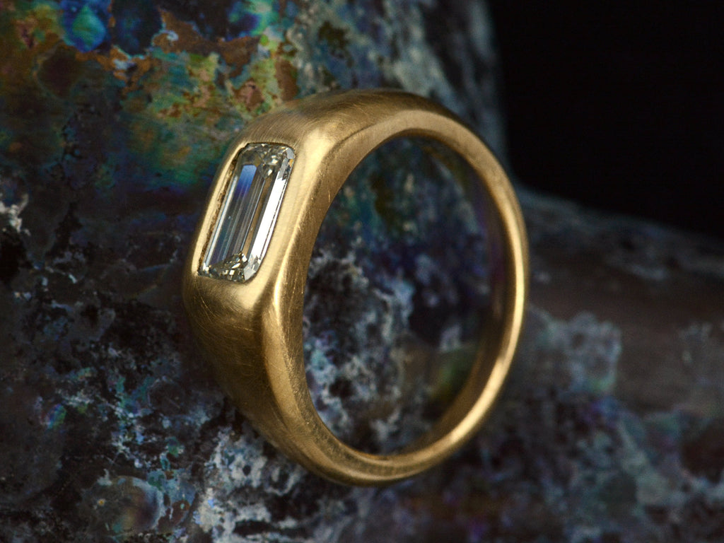 Angled side view of EB Modern engagement ring with elongated emerald cut diamond flush set in a rounded 22k yellow gold mounting, on dark background.
