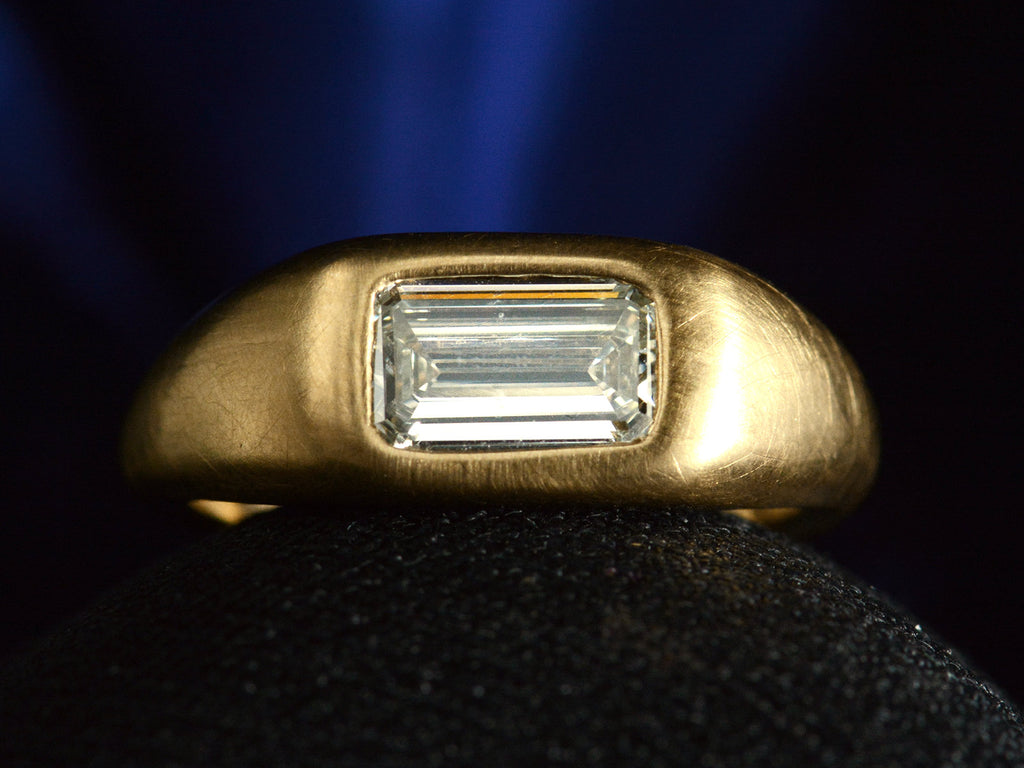 Face on view of EB Modern engagement ring with elongated emerald cut diamond flush set in a rounded 22k yellow gold mounting, on dark background.