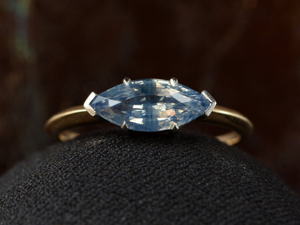 Close up front view of EB Modern ring with marquise-shaped light blue sapphire set in a simple platinum topped yellow gold mounting, shown on black domed surface.