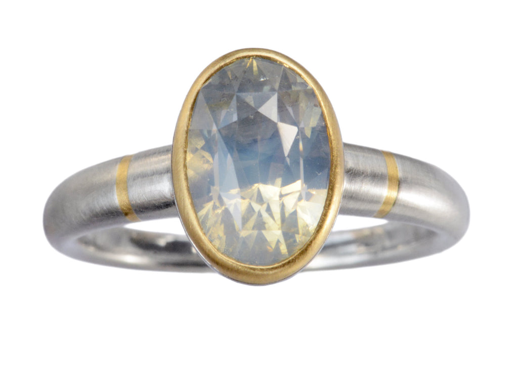 Minimal platinum ring with an oval pale blue opalescent sapphire set in a yellow gold collar with additional yellow gold stripes on the shank. Shown on white background.