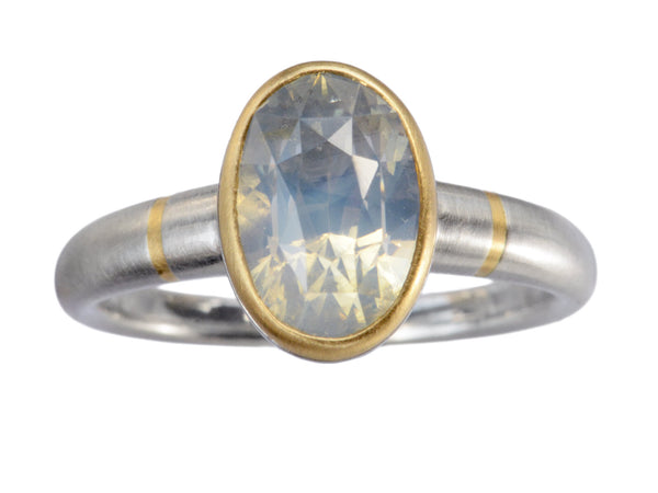 Minimal platinum ring with an oval pale blue opalescent sapphire set in a yellow gold collar with additional yellow gold stripes on the shank. Shown on white background.