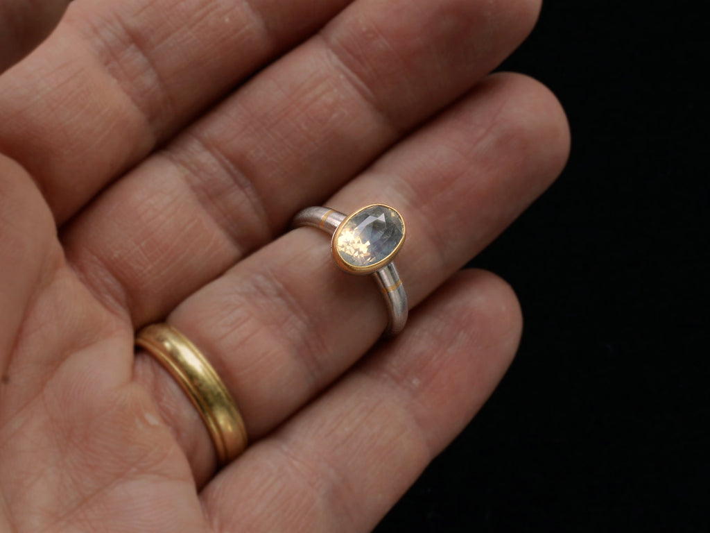 Minimal platinum ring with an oval pale blue opalescent sapphire set in a yellow gold collar with additional yellow gold stripes on the shank. Shown on hand.
