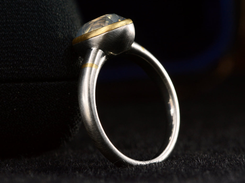 Angled side profile view of platinum ring with an oval pale blue opalescent sapphire set in a yellow gold collar with additional yellow gold stripes on the shank. Shown on dark background.