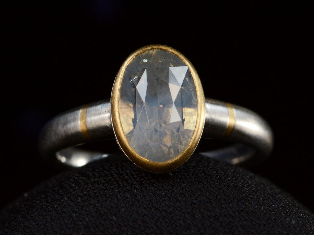 Front view of platinum ring with an oval pale blue opalescent sapphire set in a yellow gold collar with additional yellow gold stripes on the shank. Shown on dark background.