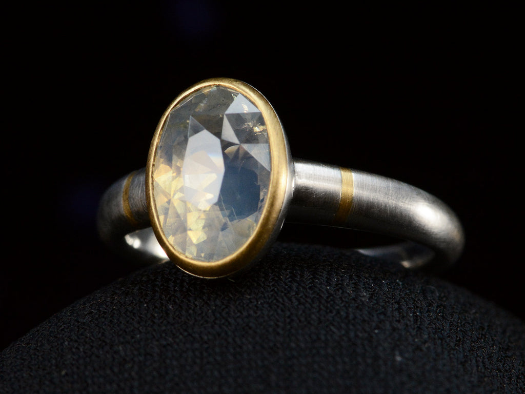 Angled front view of platinum ring with an oval pale blue opalescent sapphire set in a yellow gold collar with additional yellow gold stripes on the shank. Shown on dark background.