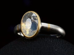 thumbnail of Angled front view of platinum ring with an oval pale blue opalescent sapphire set in a yellow gold collar with additional yellow gold stripes on the shank. Shown on dark background.