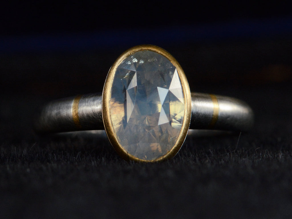 Front profile view of platinum ring with an oval pale blue opalescent sapphire set in a yellow gold collar with additional yellow gold stripes on the shank. Shown on dark background.
