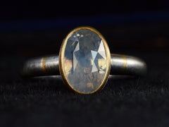 thumbnail of Front profile view of platinum ring with an oval pale blue opalescent sapphire set in a yellow gold collar with additional yellow gold stripes on the shank. Shown on dark background.