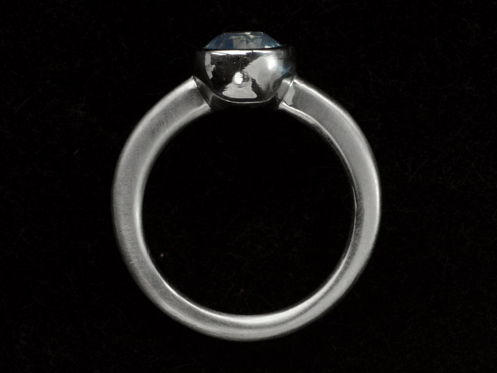 Side profile of a round opalescent light blue sapphire in a platinum bezel with a rounded matte platinum shank, shown on black background.