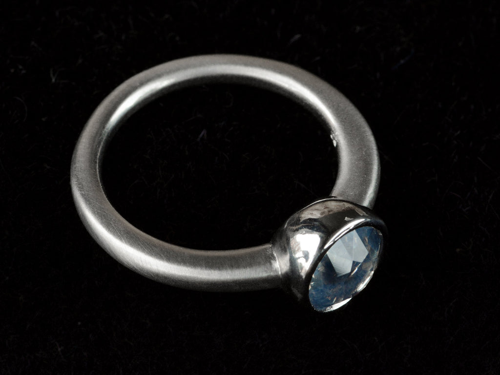 Elevated angled view of a round opalescent light blue sapphire in a platinum bezel with a rounded matte platinum shank, shown on black background.