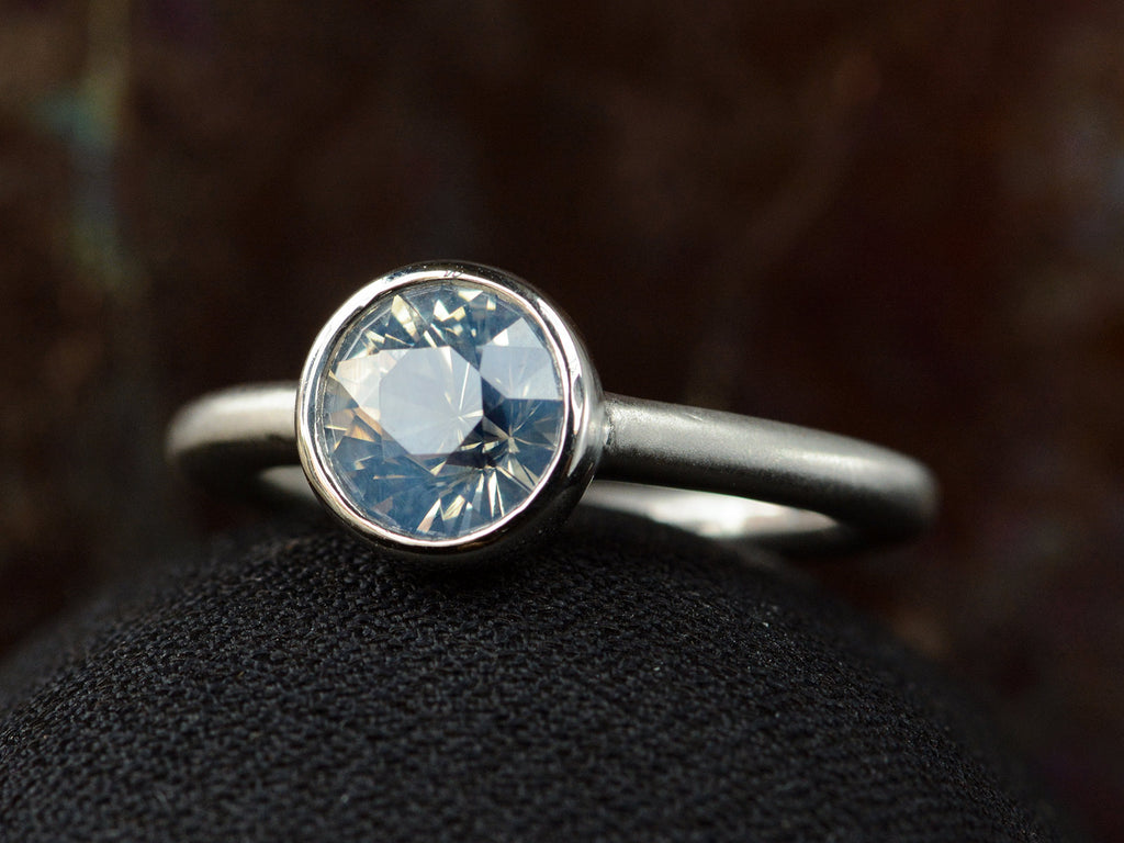 Angled front view of a round opalescent light blue sapphire in a platinum bezel with a rounded matte platinum shank, shown on dark background.