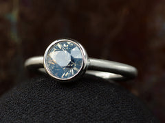 thumbnail of Angled front view of a round opalescent light blue sapphire in a platinum bezel with a rounded matte platinum shank, shown on dark background.