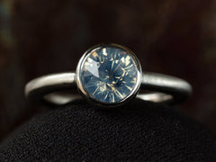 thumbnail of Front view of a round opalescent light blue sapphire in a platinum bezel with a rounded matte platinum shank, shown on dark background.