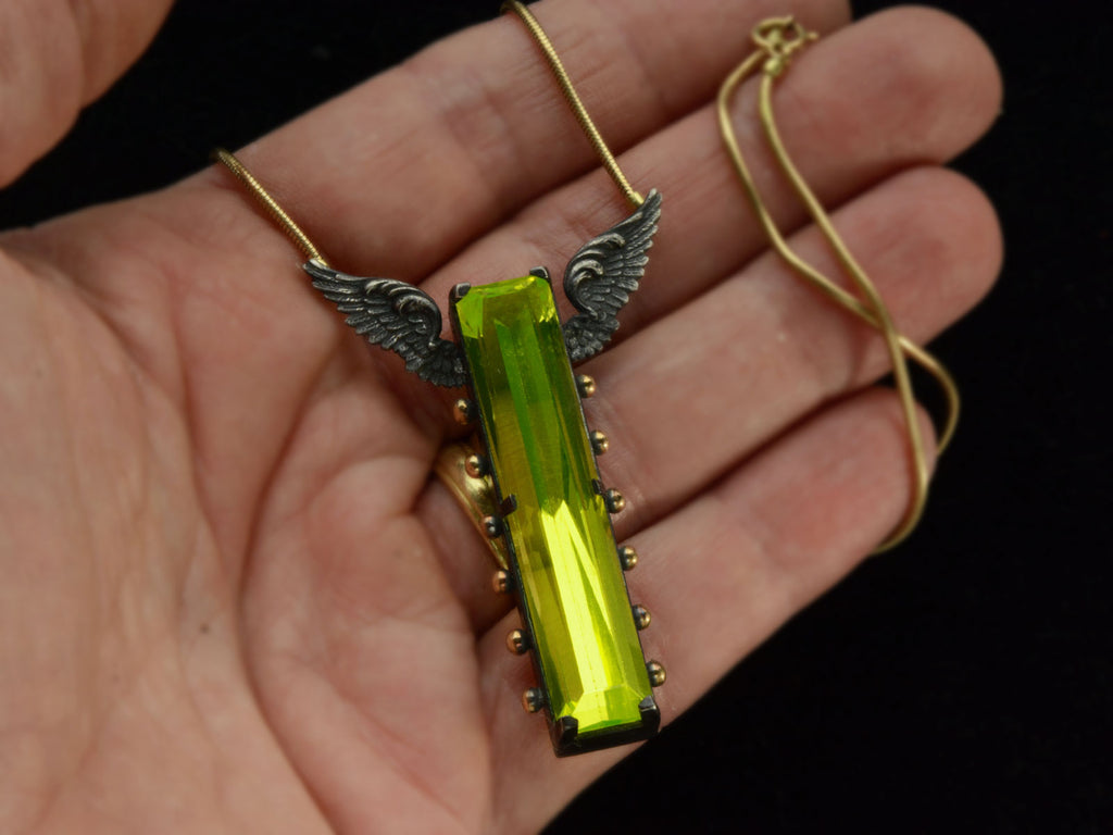 A pendant with a long florescent lime green uranium glass stone set in blackened silver with gold beads and wings on the mounting (in hand for scale)