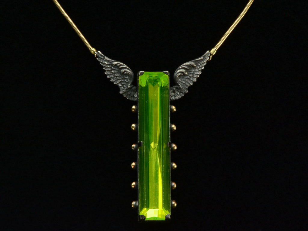 Detail of a pendant with a long florescent lime green uranium glass stone set in blackened silver with gold beads and wings on the mounting (on black background)