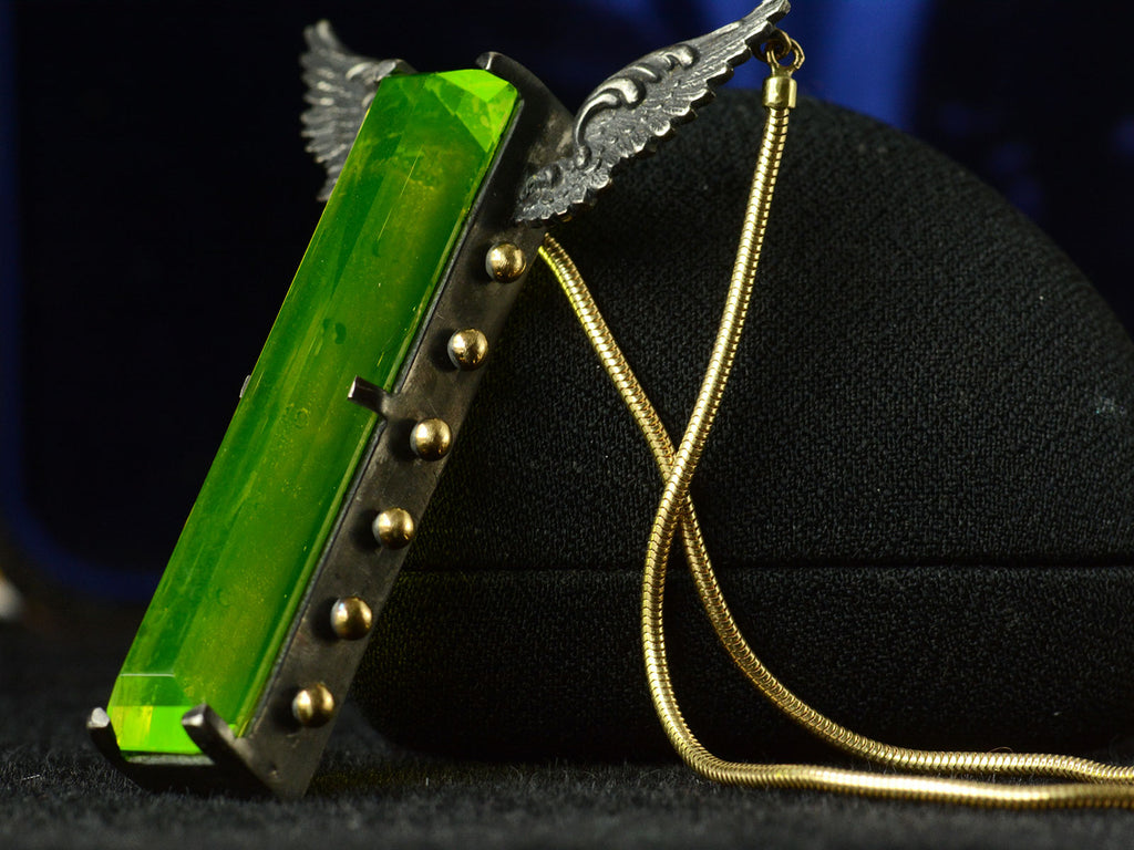 Angled view of pendant with a long florescent lime green uranium glass stone set in blackened silver with gold beads and wings on the mounting and yellow gold snake chain (on dark background)