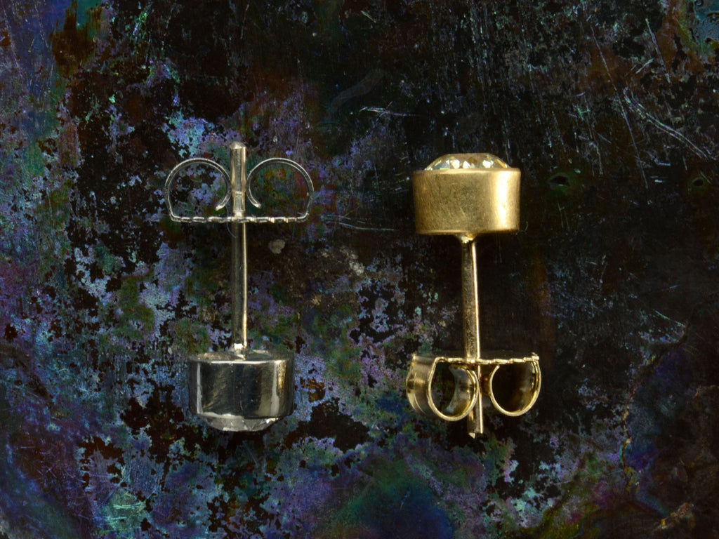 Side profile view of pair of EB Modern old European cut diamond studs, mismatched with a white diamond in platinum and a yellow diamond in 22K yellow gold. Shown on dark colorful background.