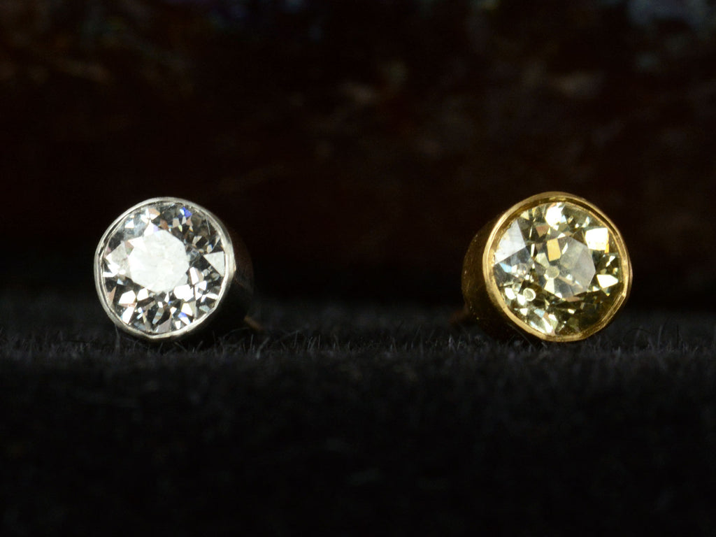 Front view of pair of EB Modern old European cut diamond studs, mismatched with a white diamond in platinum and a yellow diamond in 22K yellow gold. Shown on dark background.
