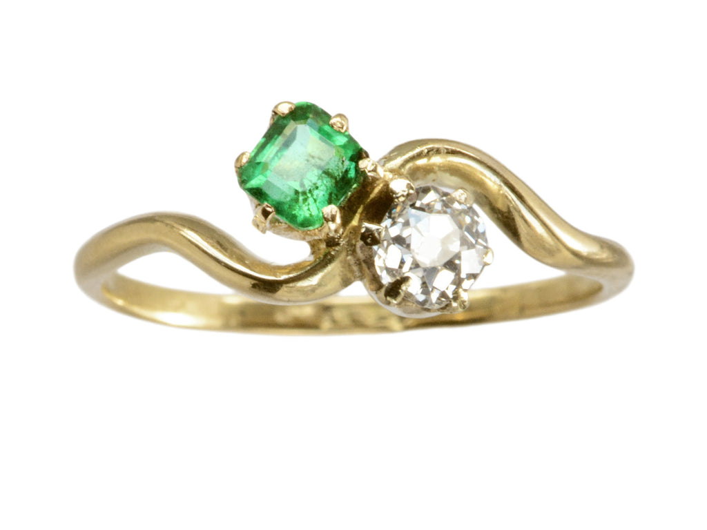 An antique Art Nouveau yellow gold wave-form ring with an emerald set opposite an old mine cut white diamond, shown on white background.