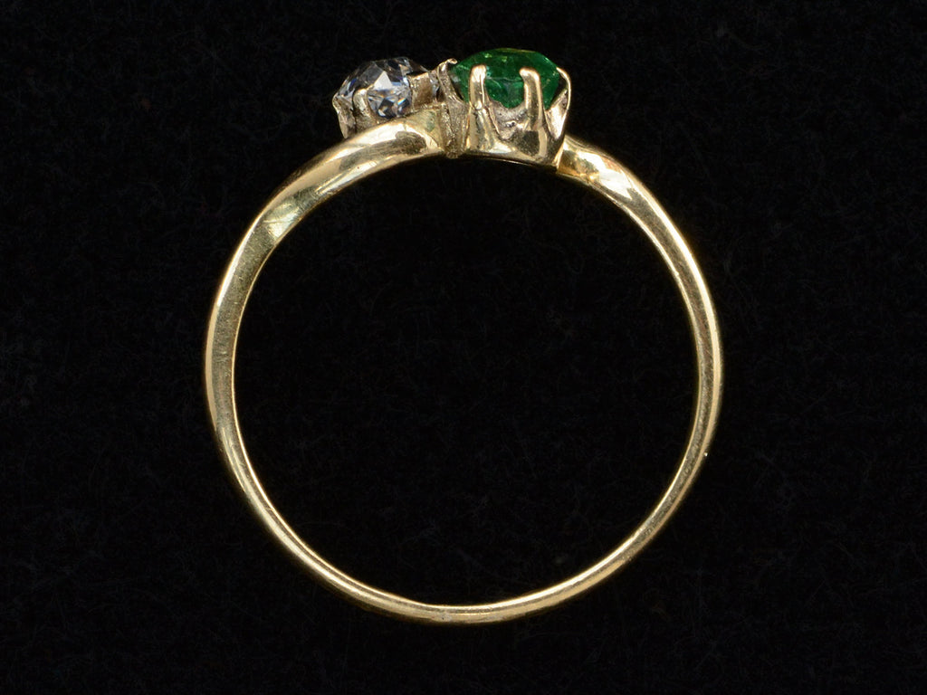 Side profile of an antique Art Nouveau yellow gold wave-form ring with an emerald set opposite an old mine cut white diamond, shown on black background.