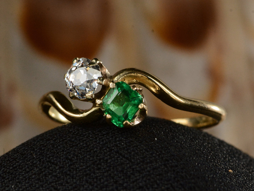Angled front view of an antique Art Nouveau yellow gold wave-form ring with an emerald set opposite an old mine cut white diamond, shown on black domed surface. 