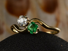 thumbnail of Angled front view of an antique Art Nouveau yellow gold wave-form ring with an emerald set opposite an old mine cut white diamond, shown on black domed surface. 