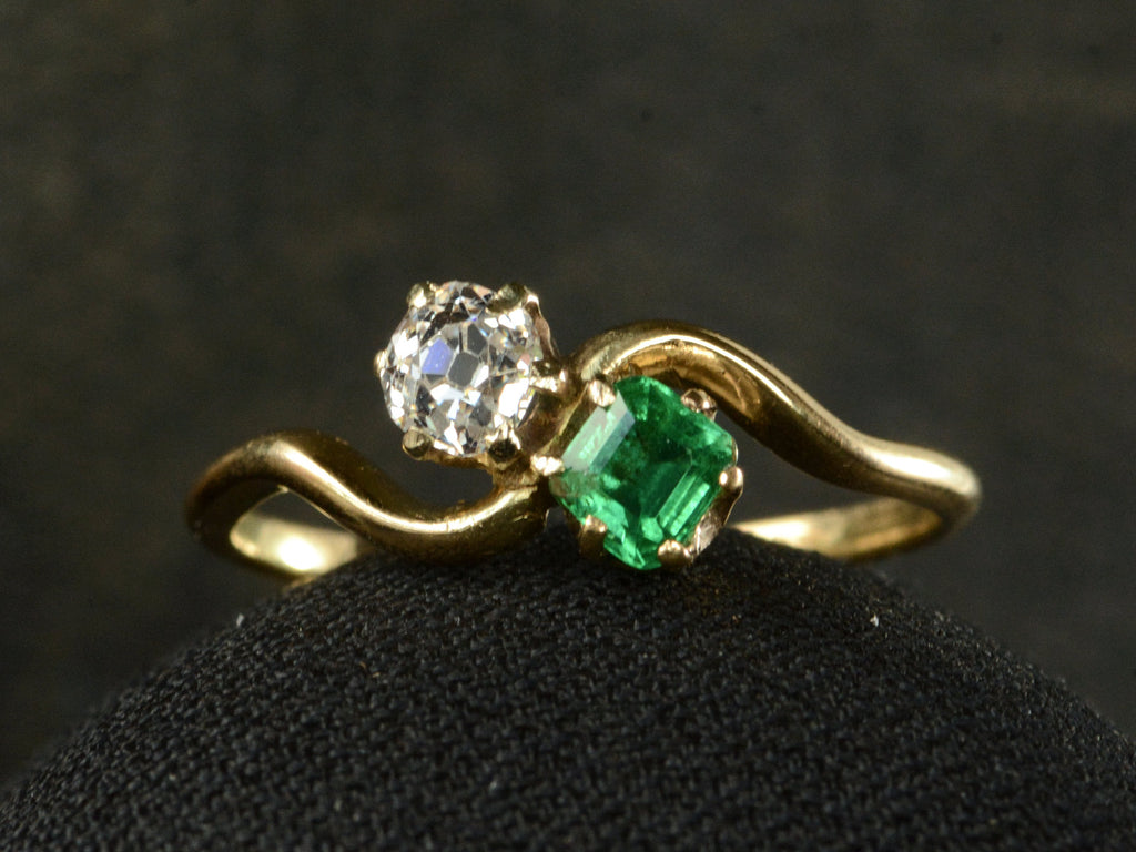 Front profile of an antique Art Nouveau yellow gold wave-form ring with an emerald set opposite an old mine cut white diamond, shown on dark background.