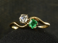 thumbnail of Front profile of an antique Art Nouveau yellow gold wave-form ring with an emerald set opposite an old mine cut white diamond, shown on dark background.
