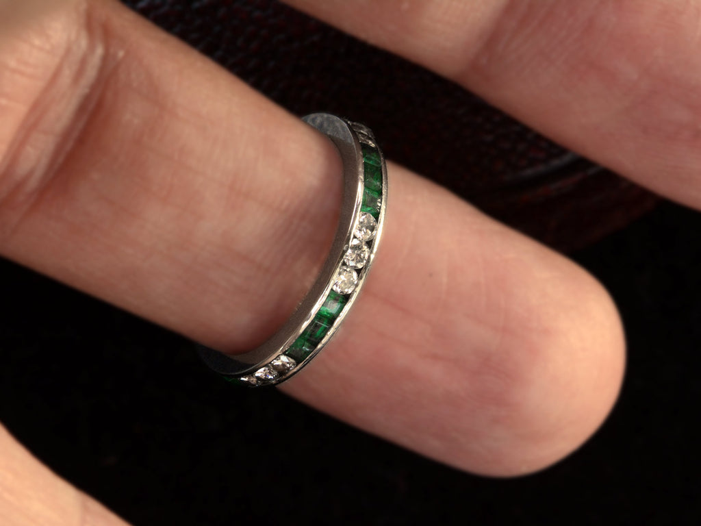 Vintage platinum eternity ring with alternating emeralds and diamonds on a finger.