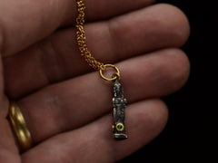 thumbnail of A small three sided figural pendant in silver and 22K yellow gold with emeralds, shown on hand.