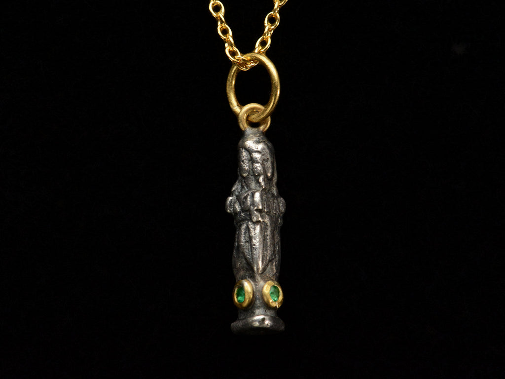 A small three sided figural pendant in silver and 22K yellow gold with emeralds, shown on dark background.