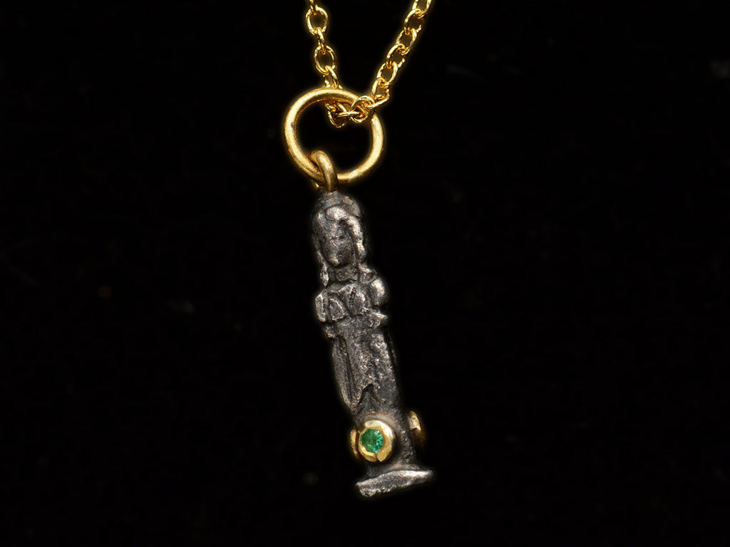Face on angle of small three sided figural pendant in silver and 22K yellow gold with emeralds, shown on dark background.