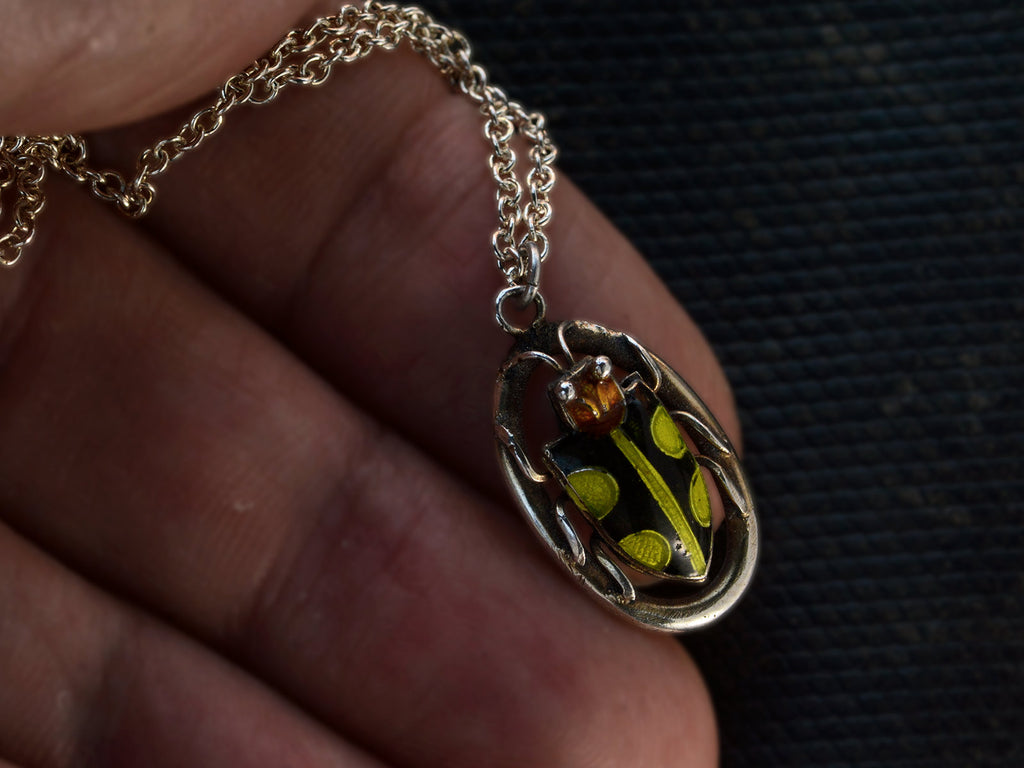 Detail of c1920 Deco silver pendant with an red-headed, black and green enamel beetle set on an oval frame, shown on held in hand.