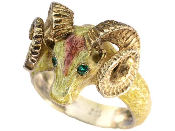 A c1960 vintage ram's head ring in yellow gold with green emerald eyes and yellow and orange enamel details. Shown on white background.