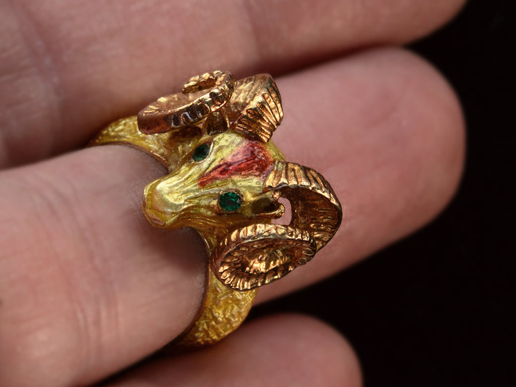 Detail view of a c1960 vintage ram's head ring in yellow gold with green emerald eyes and yellow and orange enamel details. Shown on finger.