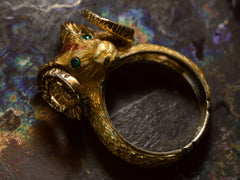 thumbnail of Angled profile view of a c1960 vintage ram's head ring in yellow gold with green emerald eyes and yellow and orange enamel details. Shown on dark colorful background.