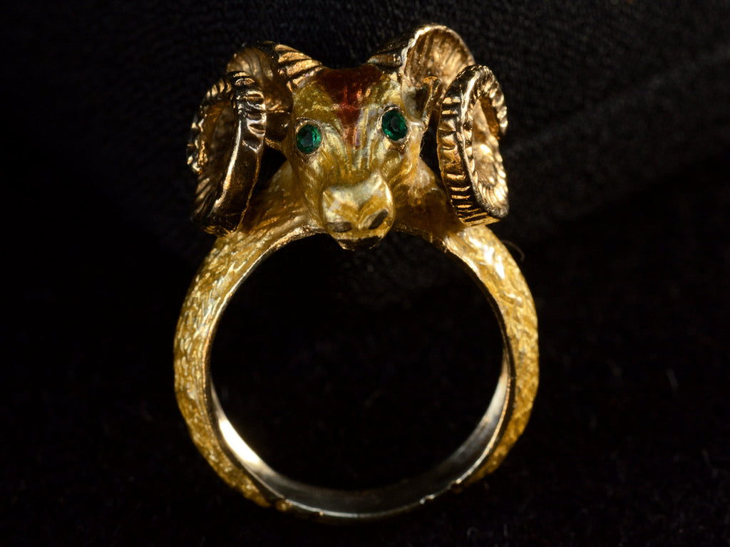 Side profile view of a c1960 vintage ram's head ring in yellow gold with green emerald eyes and yellow and orange enamel details. Shown on dark background.