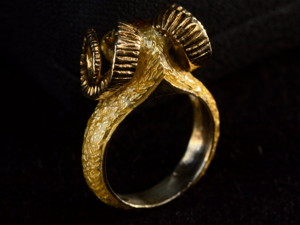 Reverse back side view of a c1960 vintage ram's head ring in yellow gold with green emerald eyes and yellow and orange enamel details. Shown on dark background.