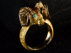 thumbnail of Elevated profile view of a c1960 vintage ram's head ring in yellow gold with green emerald eyes and yellow and orange enamel details. Shown on dark background.