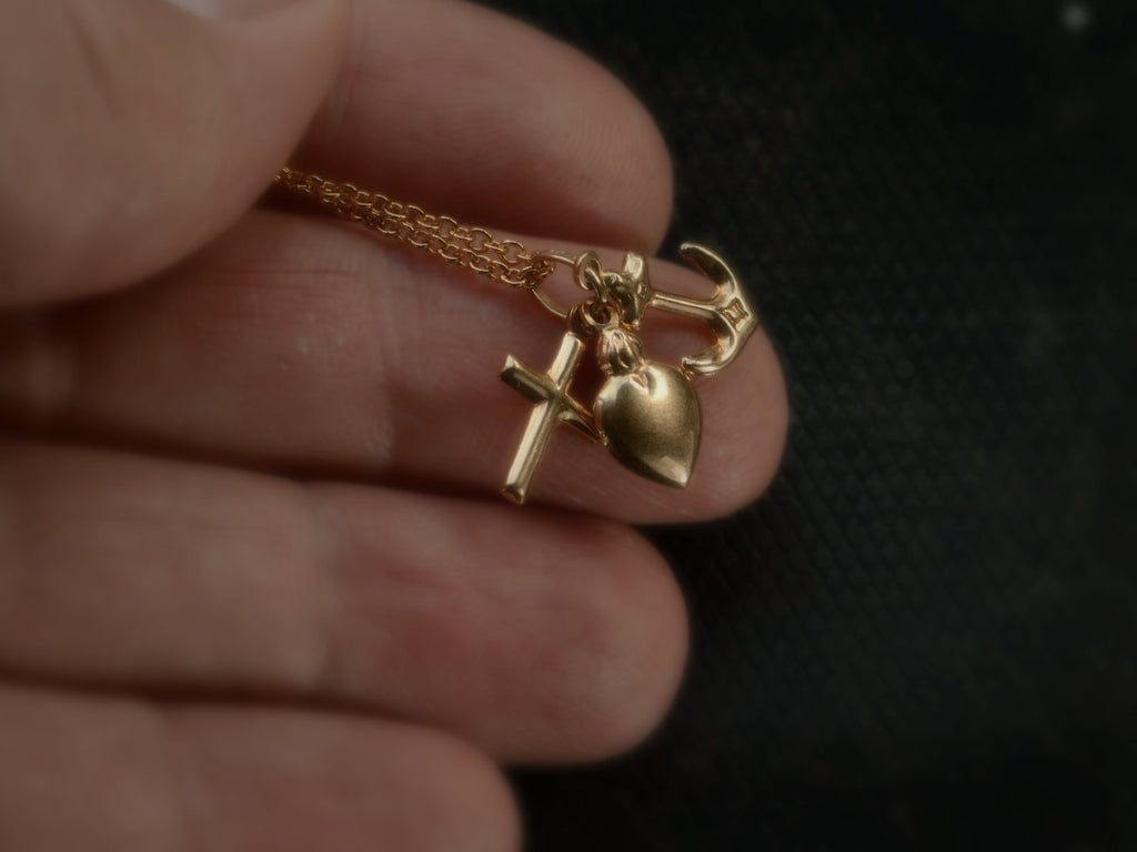 Hand holding an Italian yellow gold charm necklace with a cross, a heart and an anchor hanging on cable chain, shown on dark background.
