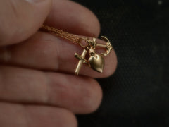 thumbnail of Hand holding an Italian yellow gold charm necklace with a cross, a heart and an anchor hanging on cable chain, shown on dark background.