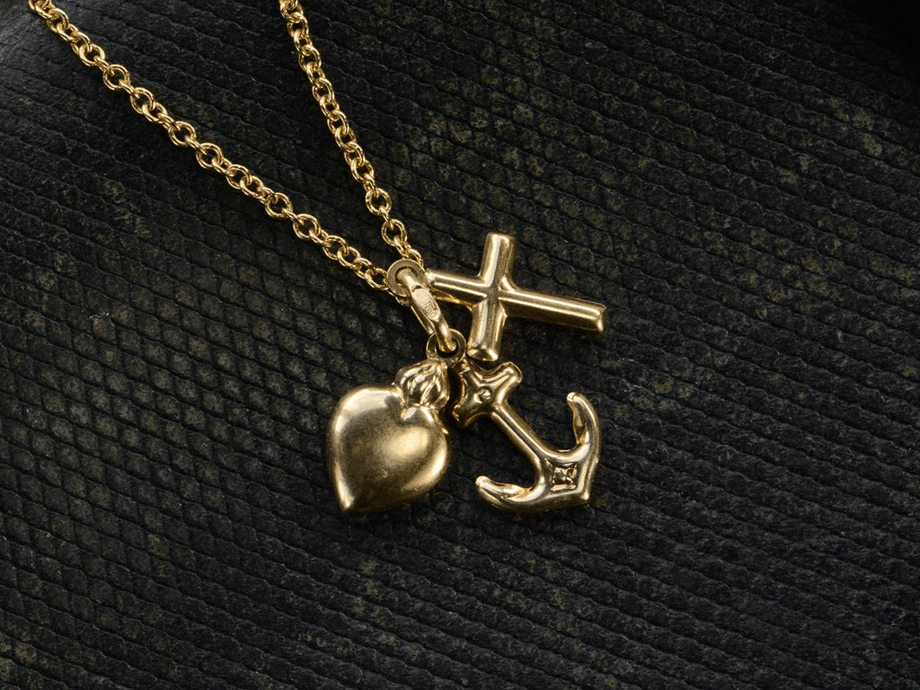 Detail of Italian yellow gold charm necklace with a cross, a heart and an anchor hanging on cable chain, shown on dark textured background.