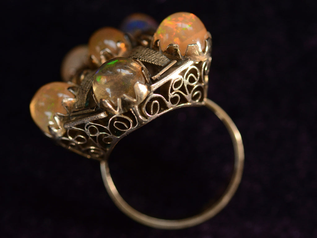 Angled profile view of pentagonal Arts & Crafts movement ring with six fire opal cabochons set in a filigreed mounting, shown on dark background.