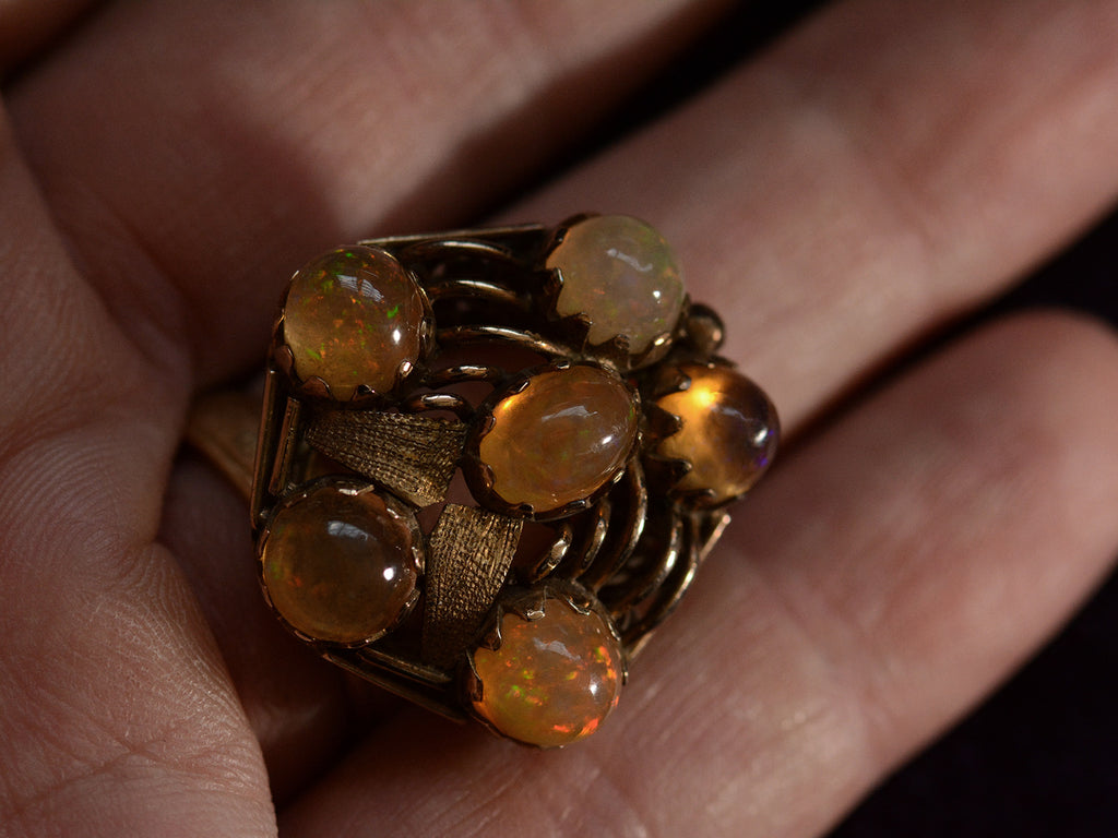 Front view of pentagonal Arts & Crafts movement ring with six fire opal cabochons set in a filigreed mounting, shown on hand for scale.