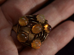 thumbnail of Front view of pentagonal Arts & Crafts movement ring with six fire opal cabochons set in a filigreed mounting, shown on hand for scale.
