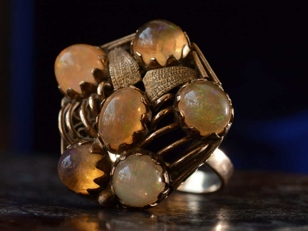 Front angled view of pentagonal Arts & Crafts movement ring with six fire opal cabochons set in a filigreed mounting, shown on dark background.