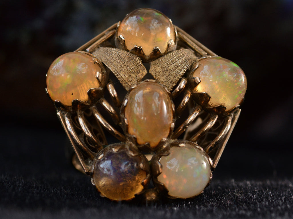 Front view of pentagonal Arts & Crafts movement ring with six fire opal cabochons set in a filigreed mounting, shown on dark background.