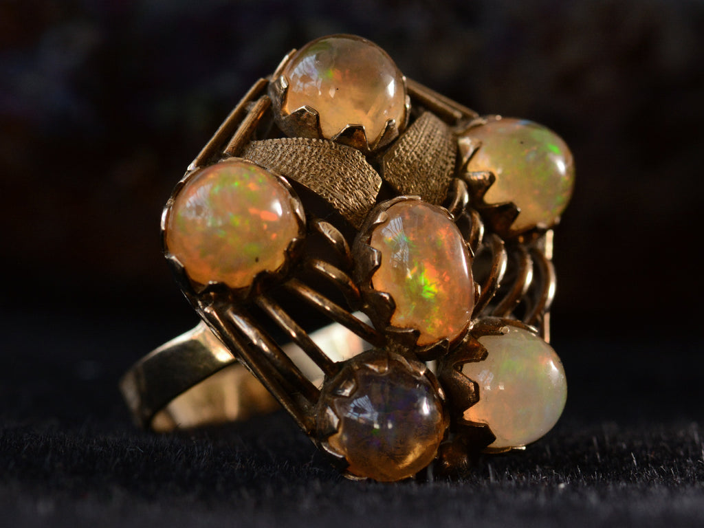 Left angled view of pentagonal Arts & Crafts movement ring with six fire opal cabochons set in a filigreed mounting, shown on dark background.